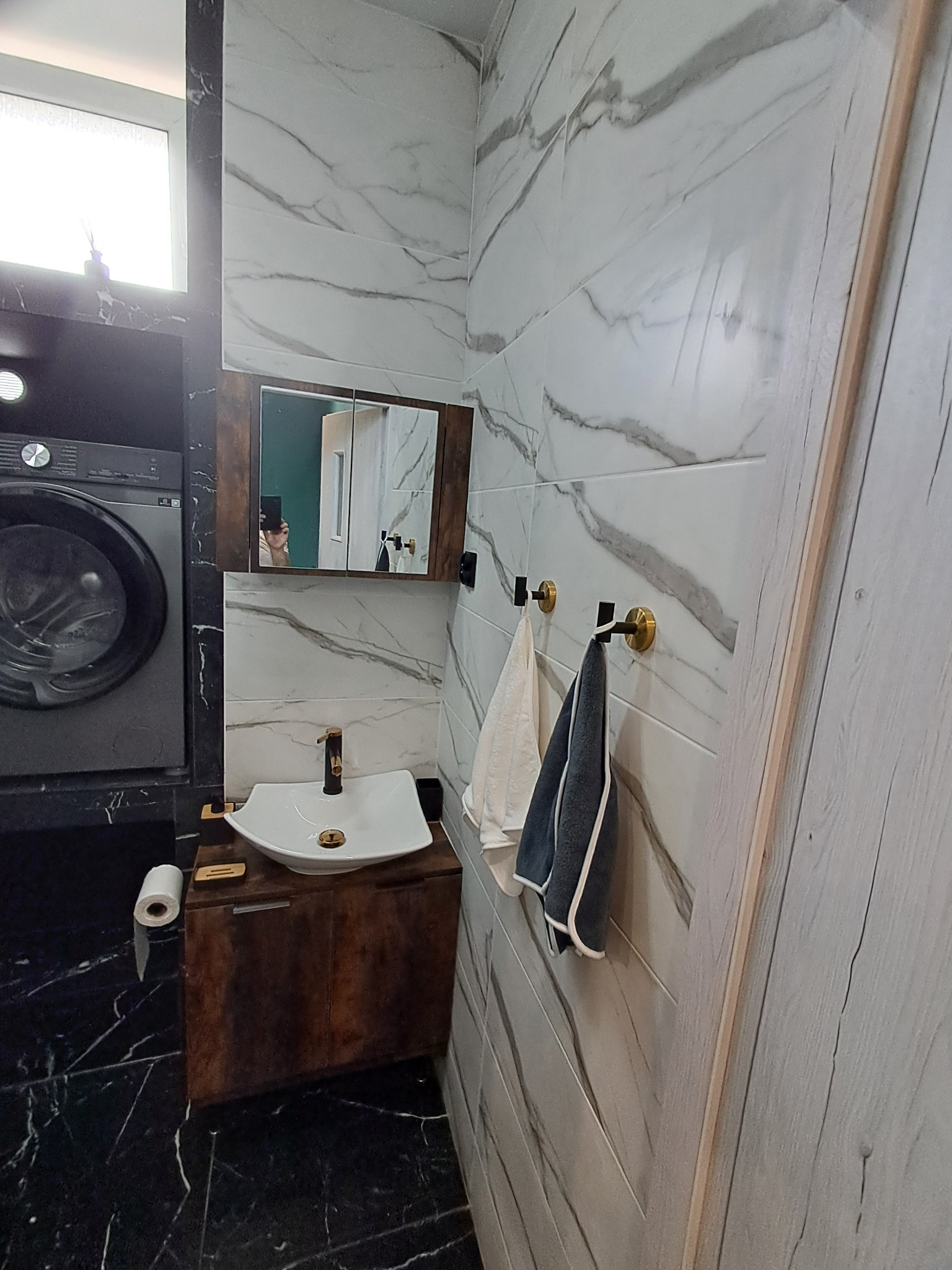 Wykończona łazienka z marmurowymi płytkami, umywalką na drewnianej szafce, pralką i lustrem. Widoczne ręczniki na złotych haczykach i rolka papieru toaletowego.