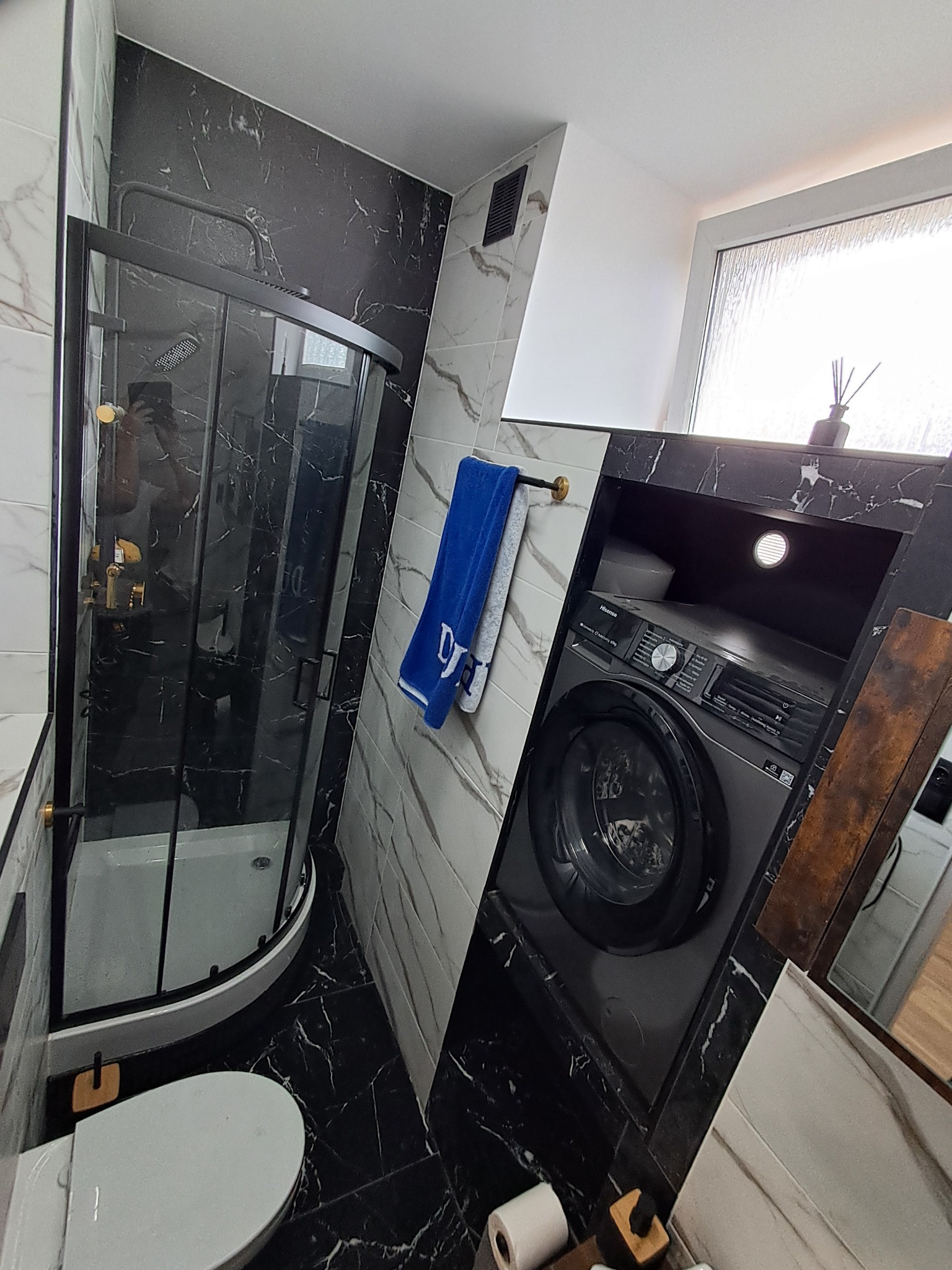 Nowoczesna, wykończona łazienka w czarno-białej kolorystyce z kabiną prysznicową, pralką w zabudowie i marmurowymi płytkami. Widok na całe wnętrze.