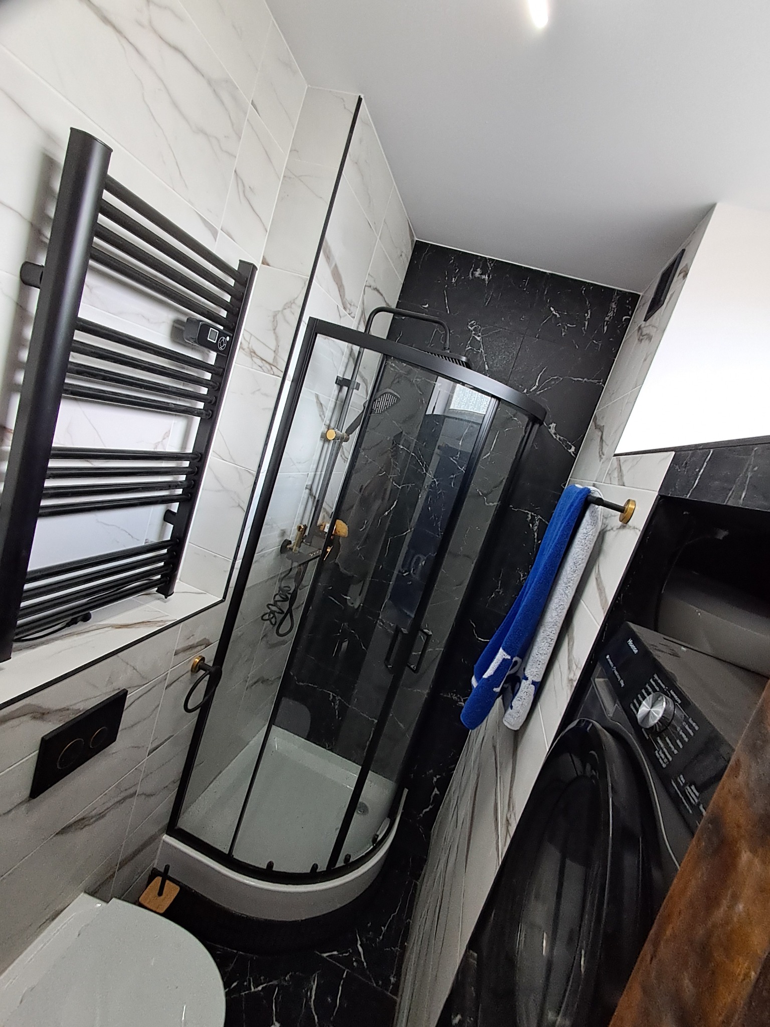 Nowoczesna łazienka z czarną kabiną prysznicową, grzejnikiem drabinkowym i pralką. Marmurowe płytki na ścianach i podłodze. Elementy wykończenia w kolorze złotym.