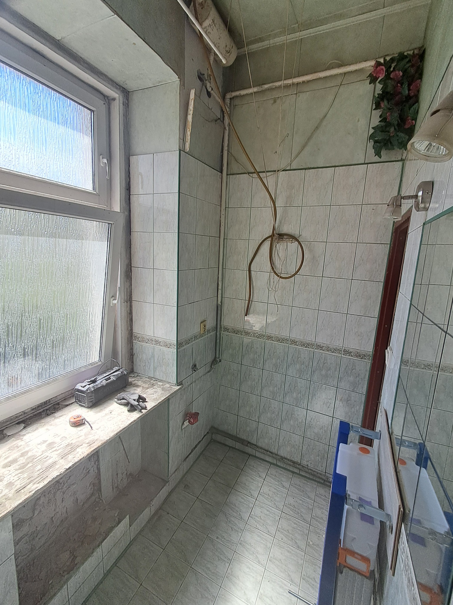 Remont łazienki w trakcie: widoczne płytki w odcieniach zieleni, okno z narzędziami na parapecie, instalacja elektryczna i częściowo zamontowana toaleta podtynkowa.