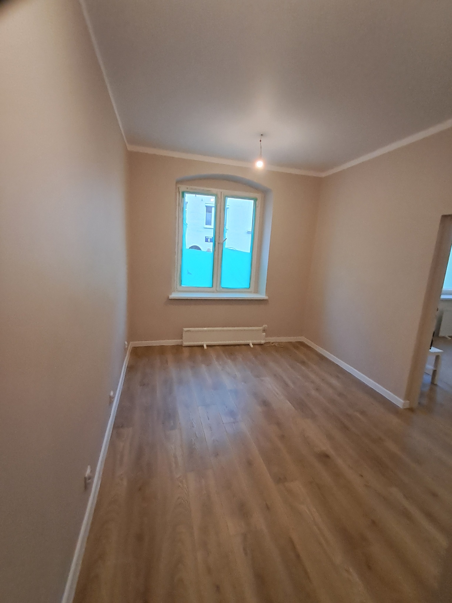 Jasne wnętrze po remoncie: świeżo pomalowane ściany w odcieniach beżu, drewnopodobne panele podłogowe, okno z niebieską folią ochronną, minimalistyczny wystrój.