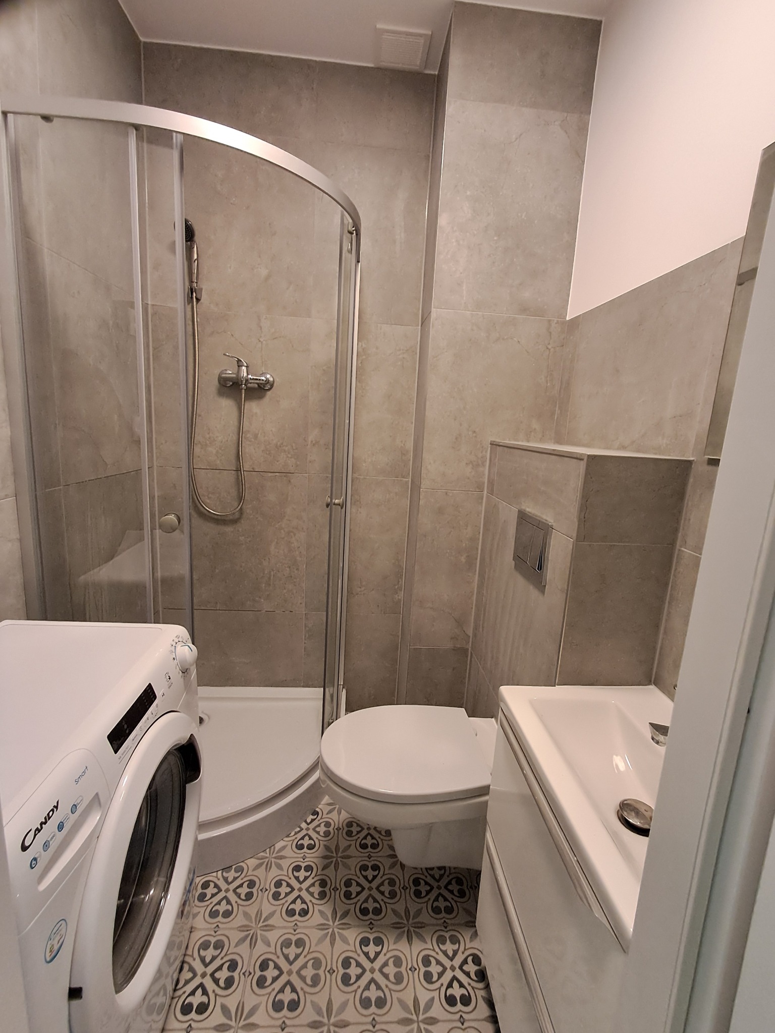 Wykończona łazienka w szarościach: kabina prysznicowa, umywalka z szafką, toaleta i pralka Candy. Wzorzysta podłoga dodaje charakteru.