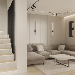 OM Dekor Sylwia Kaczmarek-Bartoszek - Jasne wnętrze salonu z narożną sofą, minimalistycznym stolikiem i schodami z podświetleniem LED. Dekoracyjny zegar na ścianie i widok na zieleń za oknem.