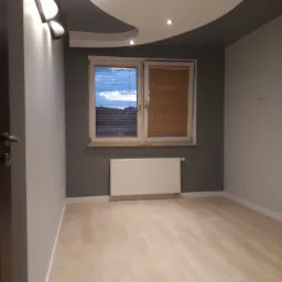 Pusty pokój po remoncie z parkietem, szarą ścianą, oknem z żaluzjami i oryginalnym oświetleniem sufitowym.