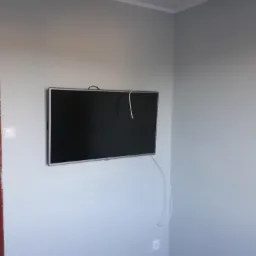Telewizor LCD zawieszony na ścianie pomalowanej na jasnoszaro, z widocznymi kablami i gniazdkiem elektrycznym w rogu pokoju.