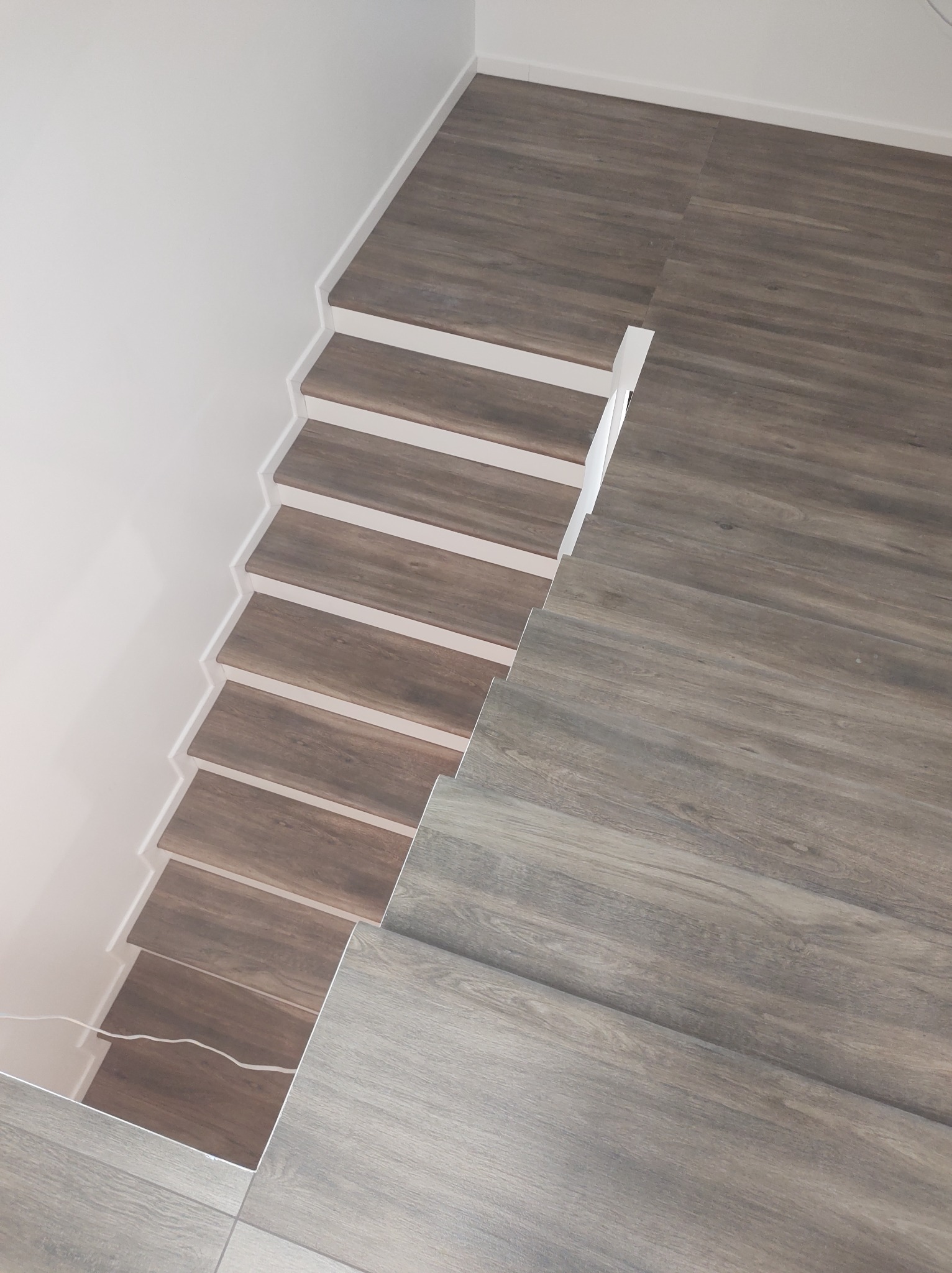 Schody wykończone panelami imitującymi drewno, z białymi podstopnicami, prowadzące na podłogę z paneli w odcieniach szarości, widok z góry.