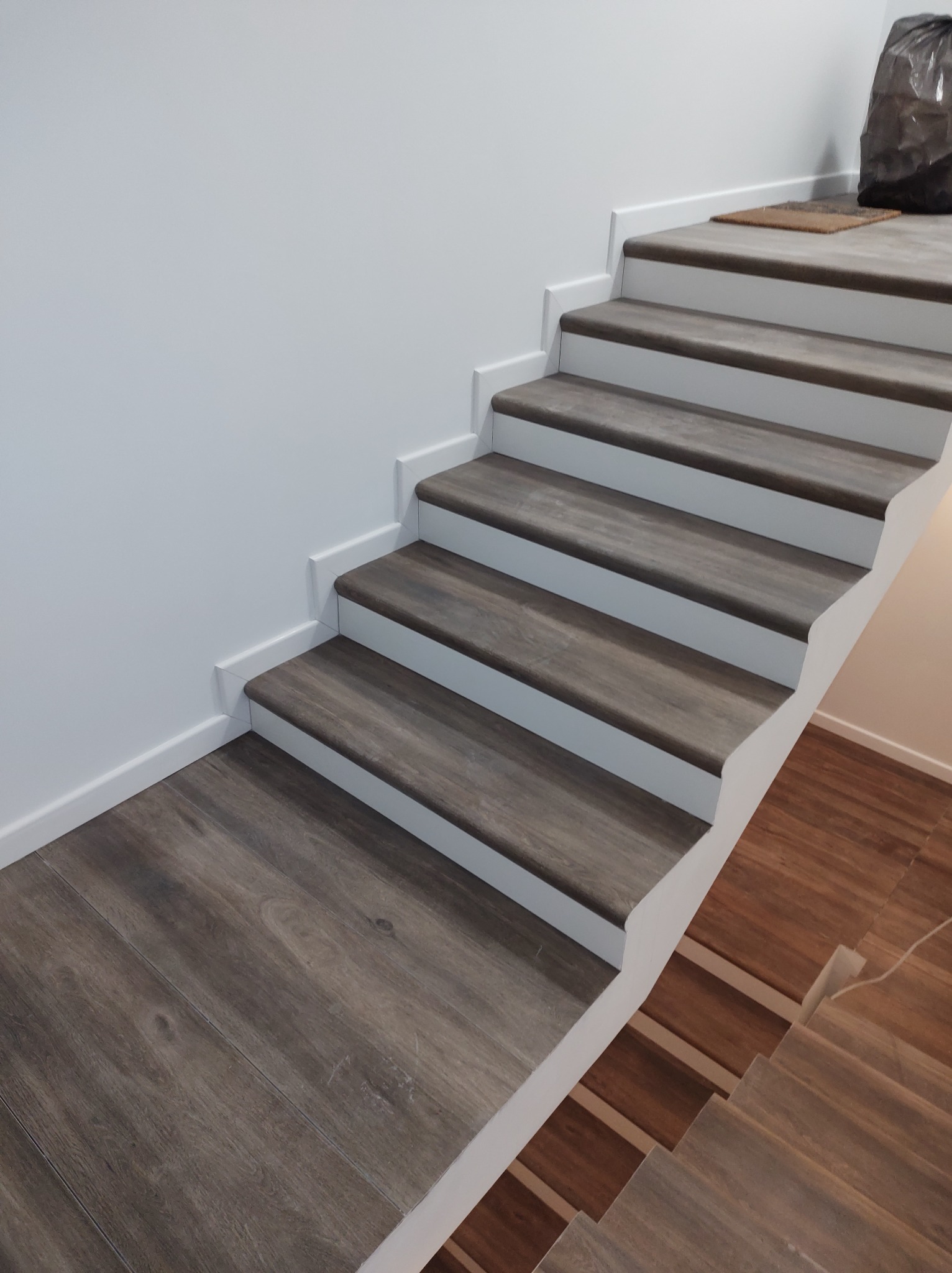 Schody wykończone panelami imitującymi drewno, z białymi listwami przypodłogowymi i obramowaniem schodów, widoczny fragment podłogi na górze schodów oraz brązowa wycieraczka i czarny worek...