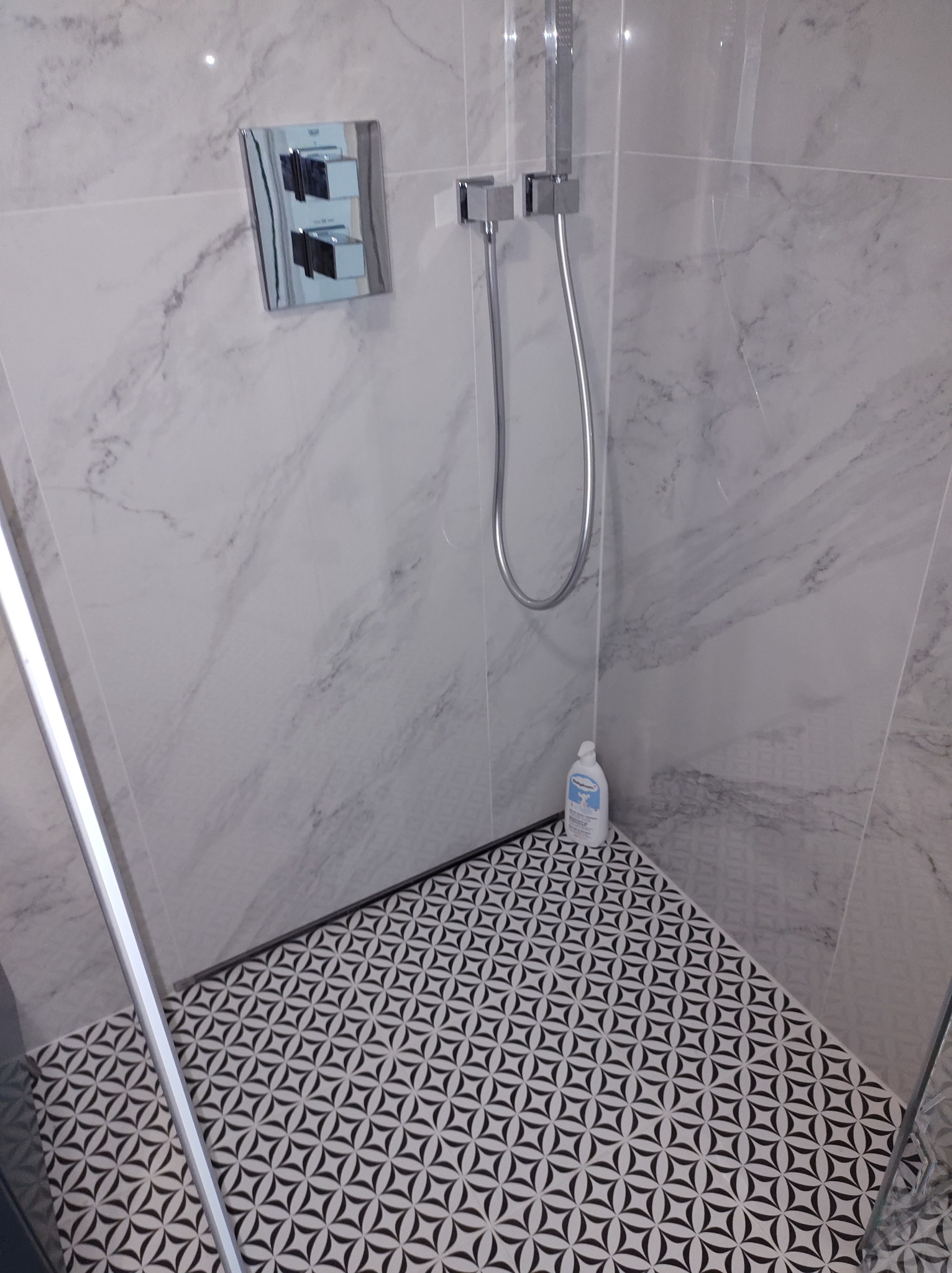 Nowoczesna kabina prysznicowa z marmurowymi płytkami ściennymi, chromowaną armaturą i geometryczną posadzką, uzupełniona butelką płynu do mycia.