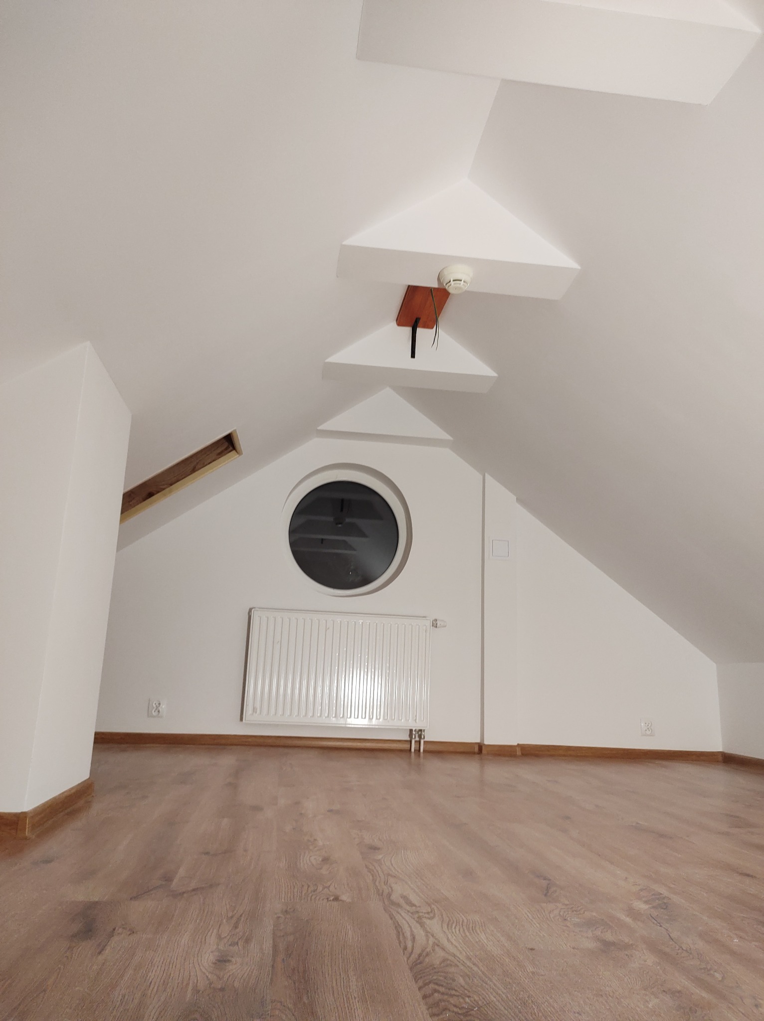 Poddasze po remoncie: białe ściany, okrągłe okno, grzejnik, drewnopodobna podłoga i czujnik dymu na suficie.