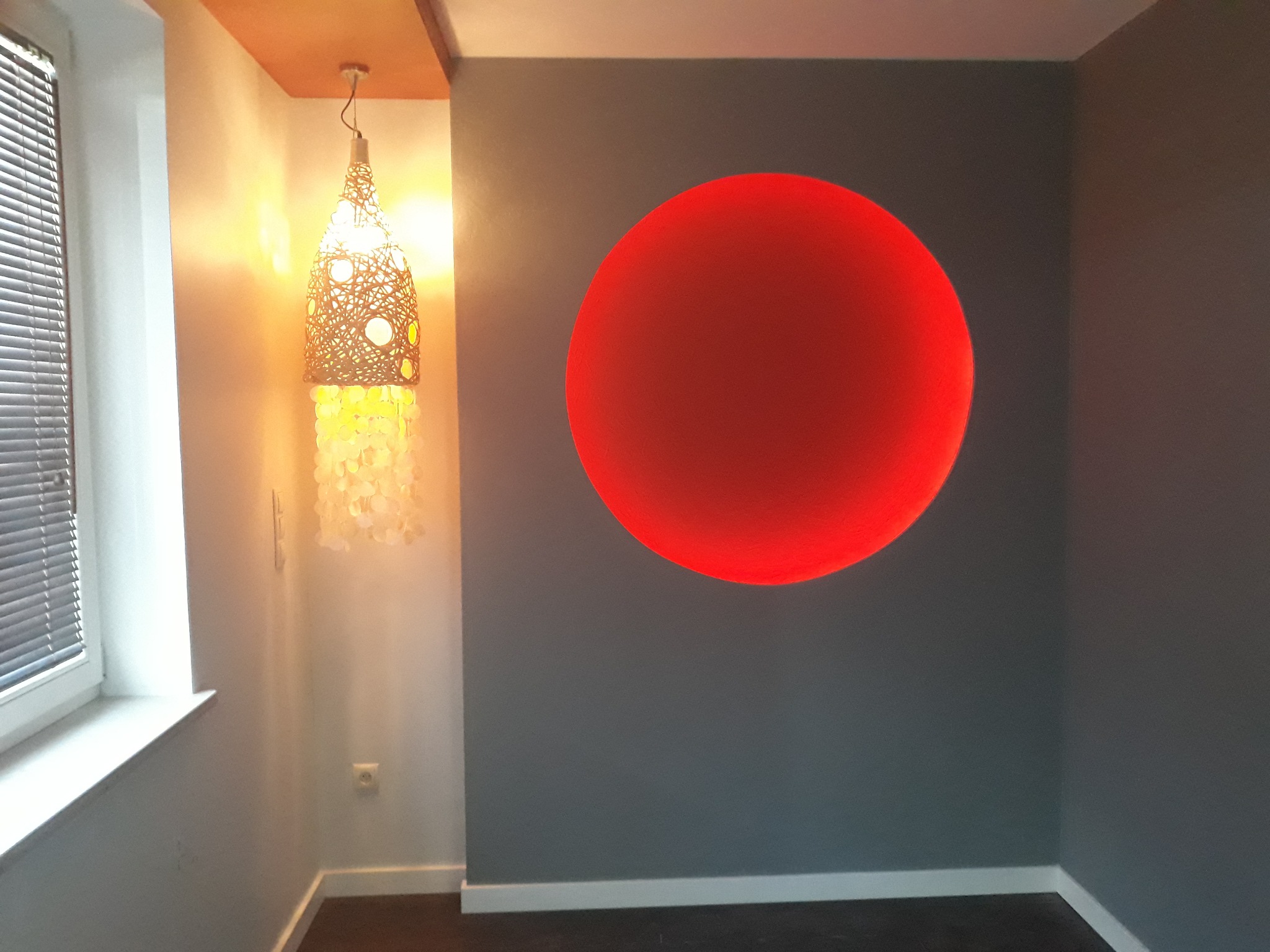 Szara ściana z namalowanym dużym, czerwonym kołem, obok okno z żaluzjami i wisząca lampa z ażurowym kloszem.