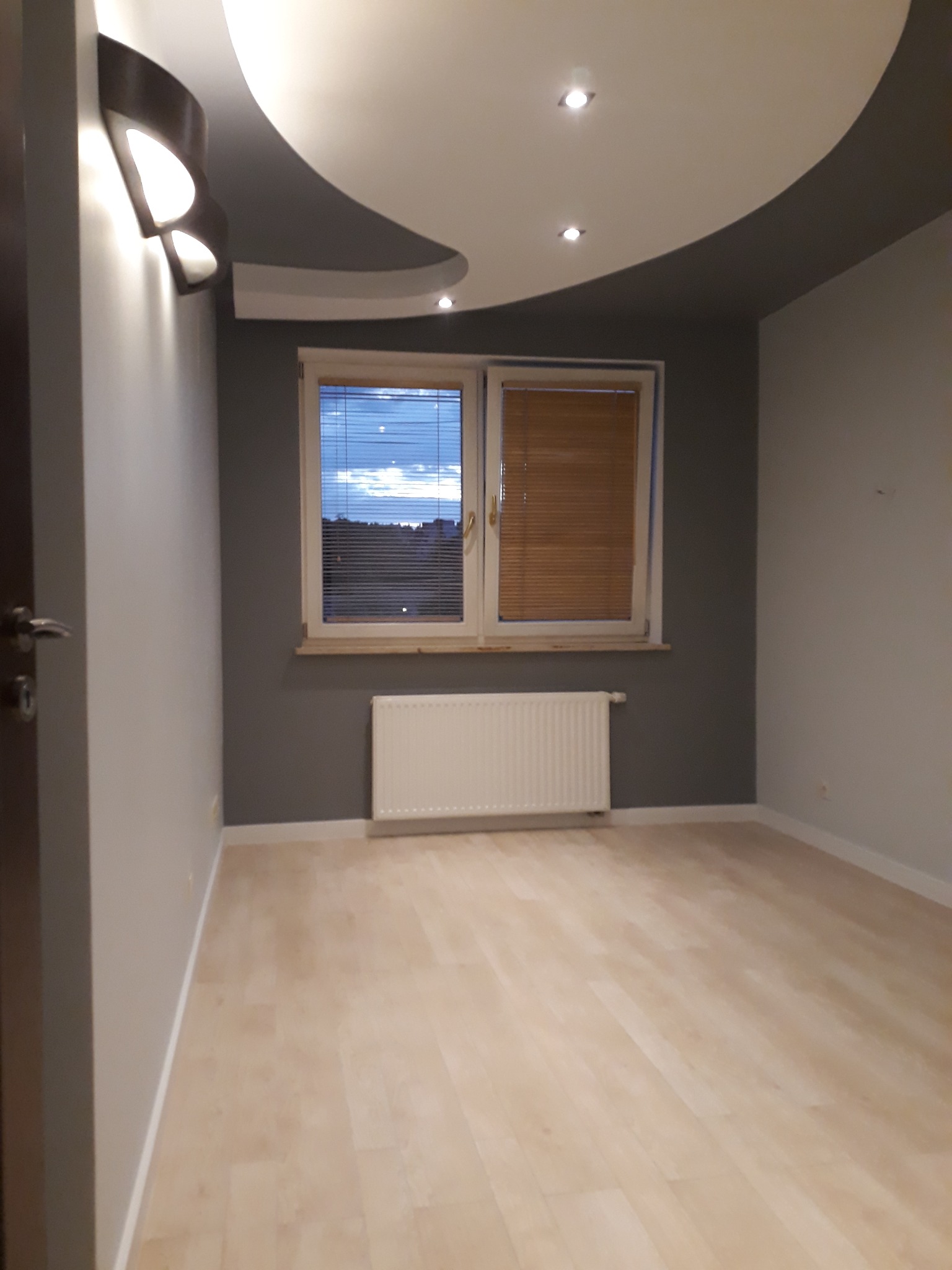 Pusty pokój po remoncie z parkietem, szarą ścianą, oknem z żaluzjami i oryginalnym oświetleniem sufitowym.