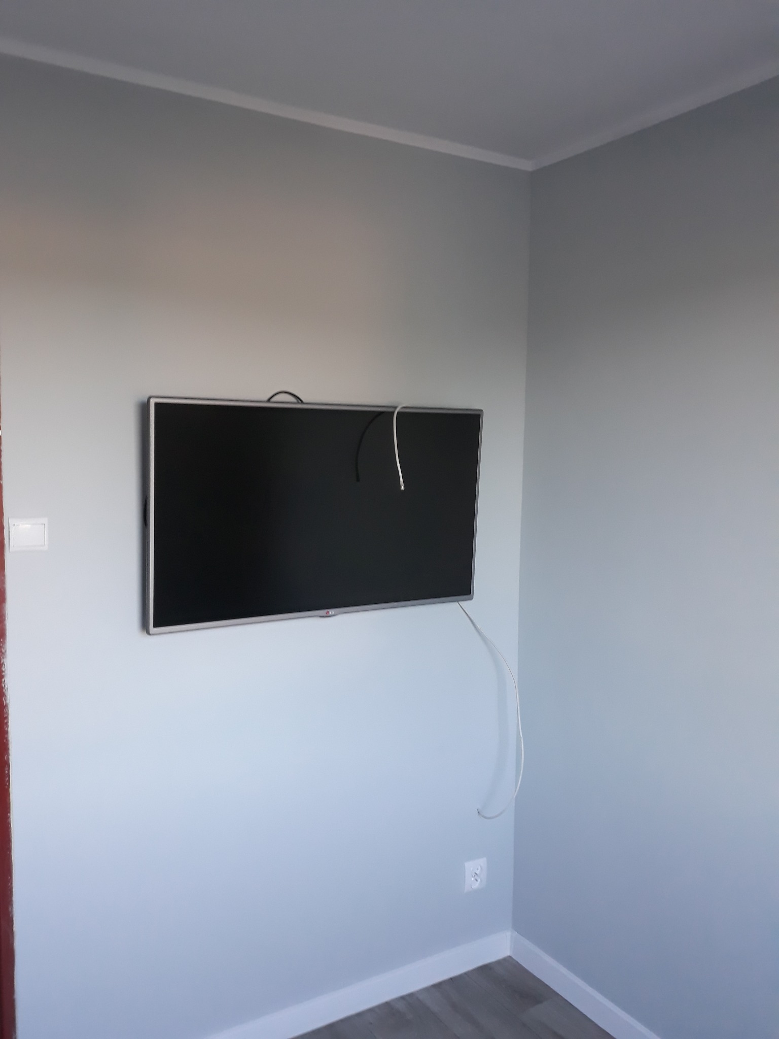 Telewizor LCD zawieszony na ścianie pomalowanej na jasnoszaro, z widocznymi kablami i gniazdkiem elektrycznym w rogu pokoju.
