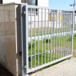 Szara brama przesuwna z pionowymi szczeblami, zamontowana na kamiennym słupku, z widocznym mechanizmem automatycznego otwierania, na tle trawnika i ogrodzenia z siatki.