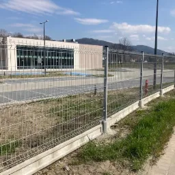 Szare ogrodzenie z paneli siatkowych na betonowej podmurówce, zabezpieczające teren budynku z parkingiem, widoczne góry w tle.