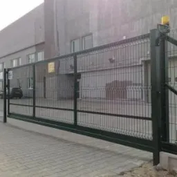 Przesuwna brama przemysłowa z wypełnieniem z siatki zgrzewanej, z żółtą lampą ostrzegawczą, na tle szarego budynku przemysłowego.