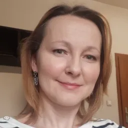 Portret uśmiechniętej kobiety z brązowymi oczami i kolczykami w geometryczne wzory, ubrana w bluzkę w granatowe paski.