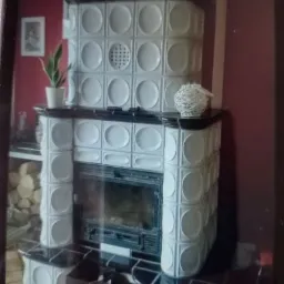 Kafelkowy piec kominkowy z białymi kaflami i czarnymi akcentami, dekoracje na górze, drewno obok, czerwona ściana w tle.