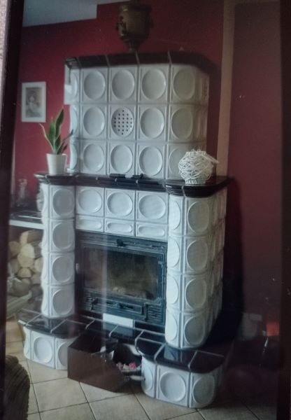 Kafelkowy piec kominkowy z białymi kaflami i czarnymi akcentami, dekoracje na górze, drewno obok, czerwona ściana w tle.