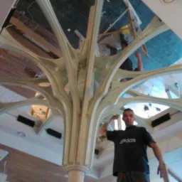 Pracownik na rusztowaniu prezentuje skomplikowaną, gipsową konstrukcję sufitową w stylu gotyckim, z widocznymi elementami instalacji elektrycznej w tle.