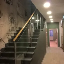 Klatka schodowa z czarnymi płytkami na stopniach i szklaną balustradą, ściana zdobiona rysunkami w stylu Leonarda da Vinci, oświetlenie sufitowe punktowe.