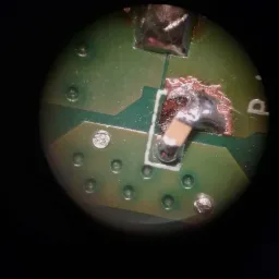 Zbliżenie na naprawiony element elektroniczny na zielonej płytce drukowanej, widoczne punkty lutownicze i ślady topnika, całość sfotografowana pod dużym powiększeniem.