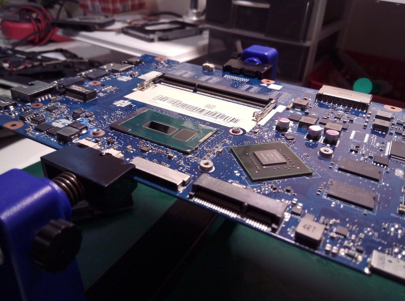 Płyta główna laptopa zamocowana w uchwycie serwisowym, widoczne układy scalone, procesor i złącza, przygotowana do naprawy lub wymiany komponentów.