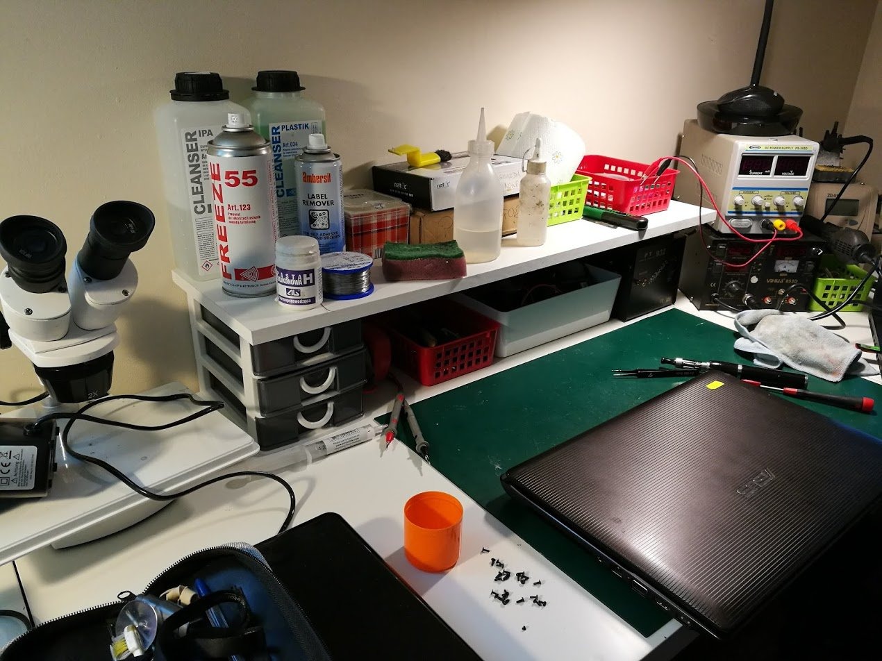 Stanowisko pracy z rozłożonym laptopem, narzędziami, mikroskopem i preparatami czyszczącymi. Widoczne śrubki, pensety, zasilacz laboratoryjny oraz preparaty chemiczne w pojemnikach. Ujęcie z bliska...