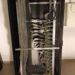 Szafa serwerowa z uporządkowanymi kablami sieciowymi, widoczne panele krosowe i przewody w kolorze szarym i żółtym, montowane na metalowych szynach wewnątrz czarnej obudowy.