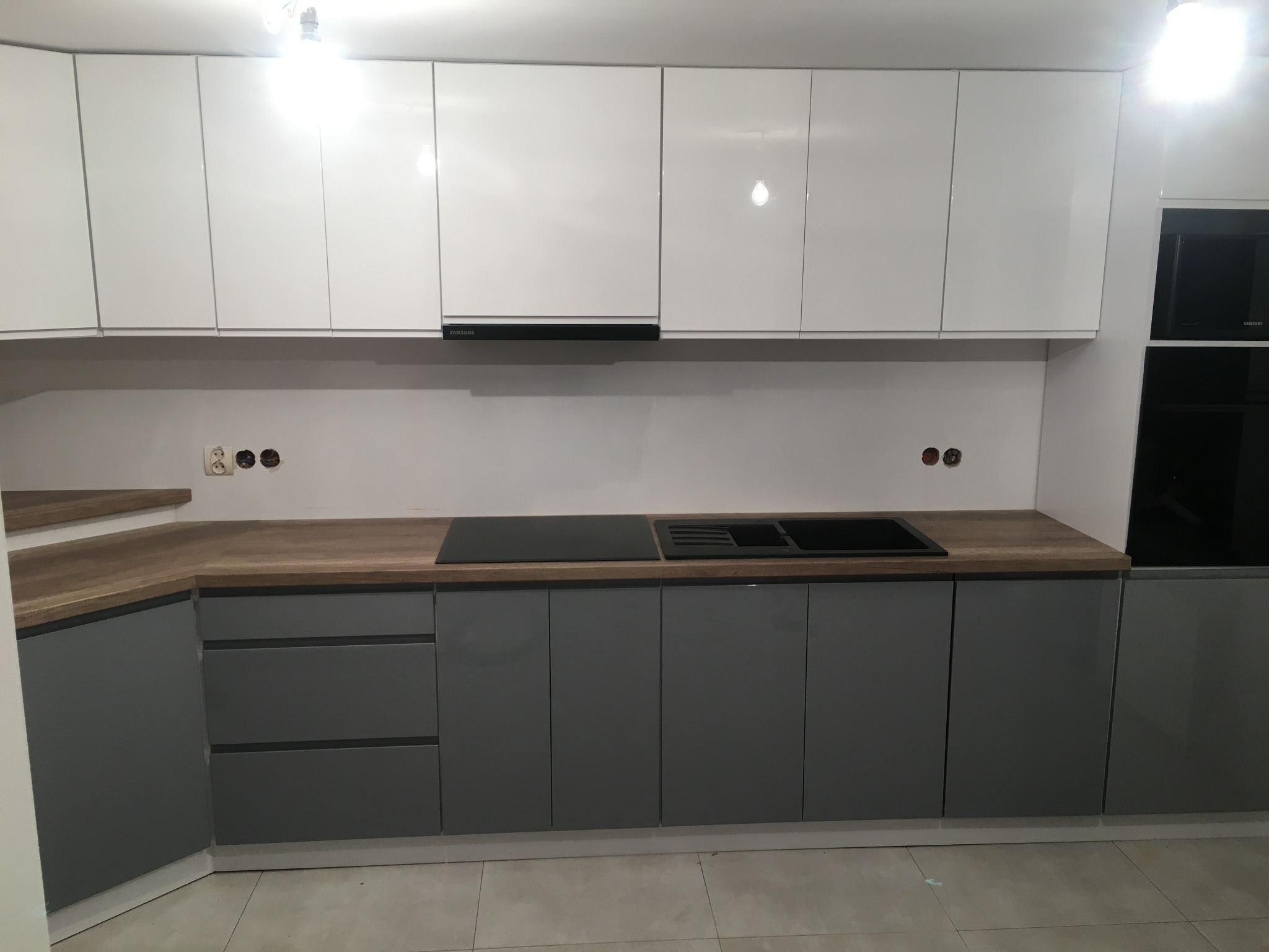 Nowoczesna kuchnia z szarymi szafkami dolnymi i białymi górnymi, drewniany blat, czarna płyta indukcyjna i zlew, widoczne gniazdka elektryczne.