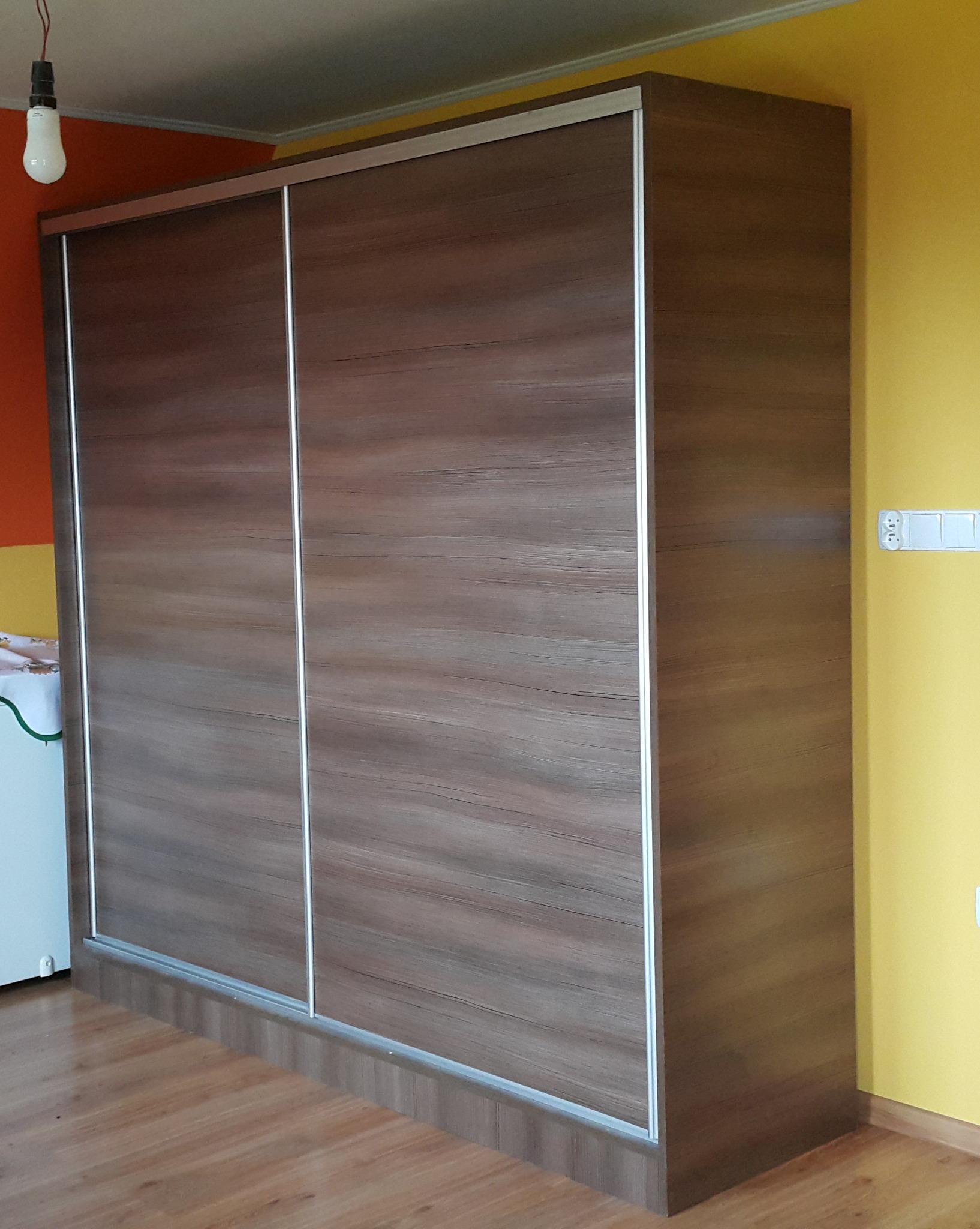 Duża szafa przesuwna z drewnopodobnymi frontami i srebrnymi obramowaniami, wykonana na wymiar w Dębnie, stoi na drewnianej podłodze, a w tle widać pomarańczową i żółtą ścianę.