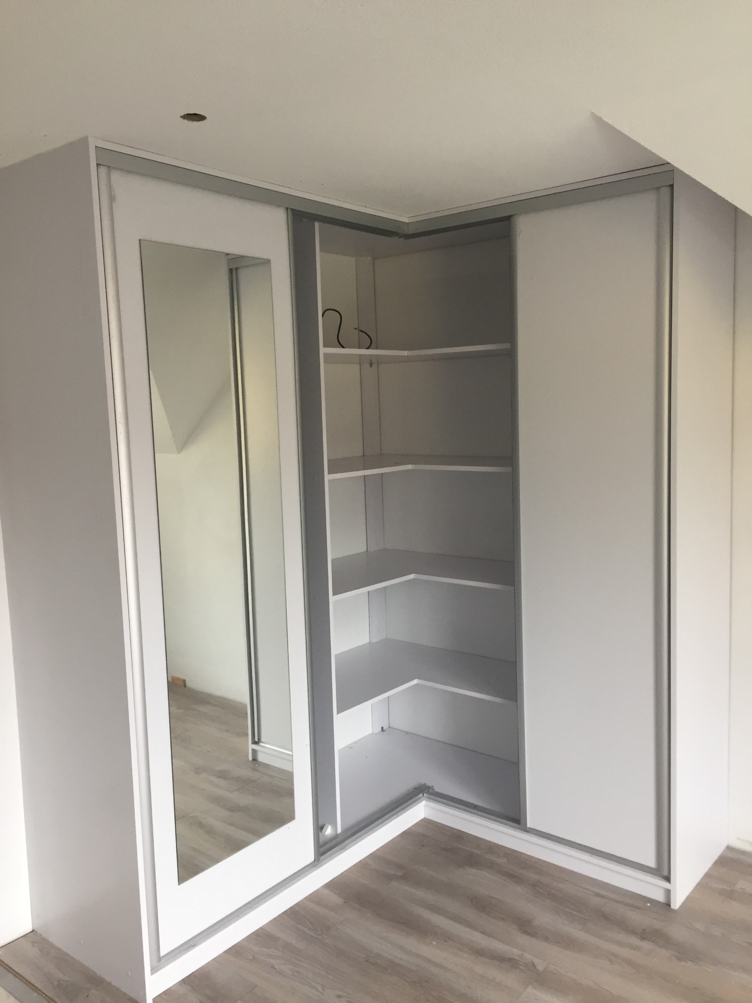 Biała szafa narożna na wymiar z przesuwnymi drzwiami, jedna z nich z lustrem, półki wewnętrzne widoczne, dopasowana do skosu dachu.