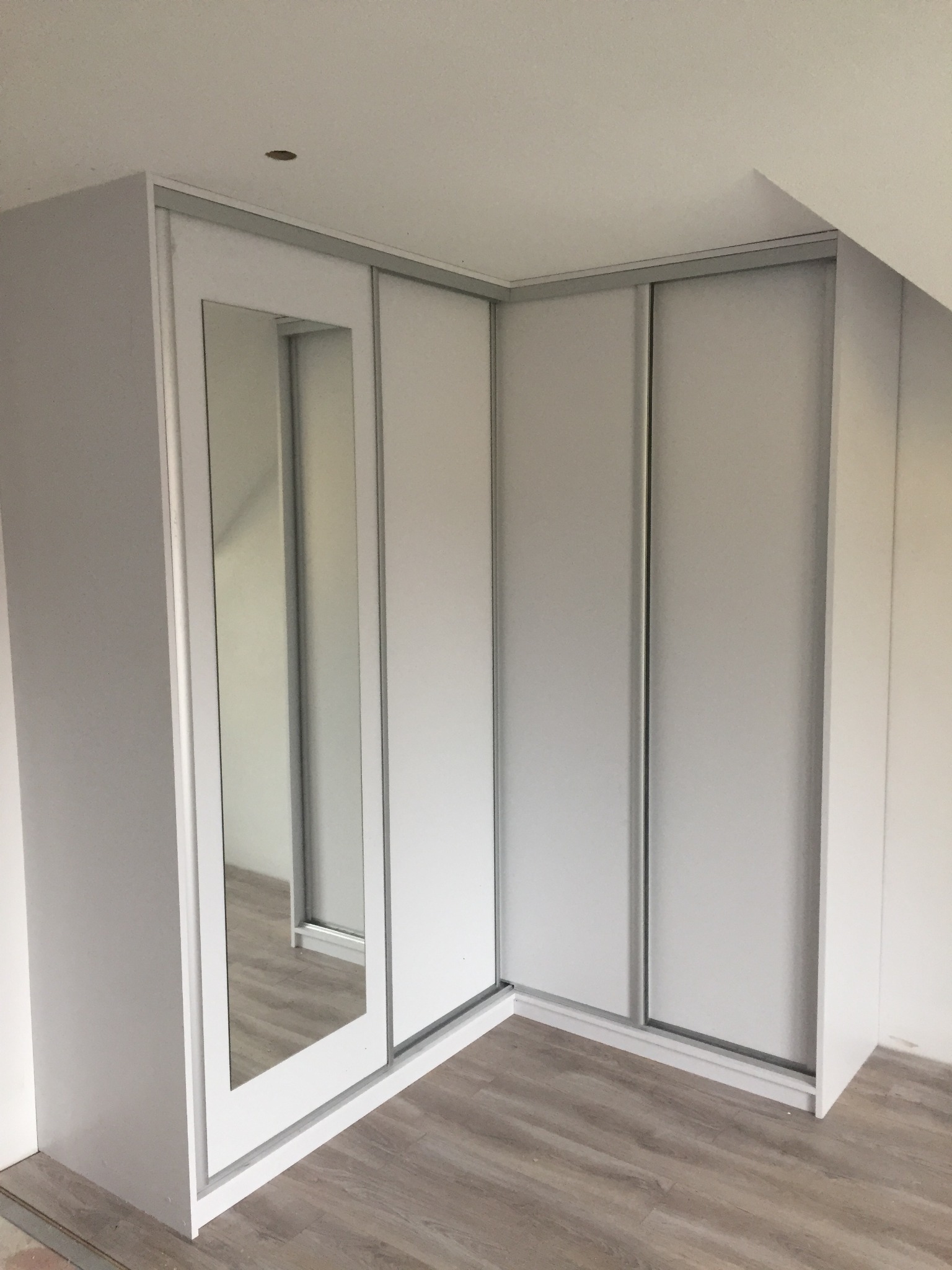Biała, narożna szafa wnękowa z przesuwnymi drzwiami, w jednym skrzydle lustro, zamontowana na poddaszu ze skosem.