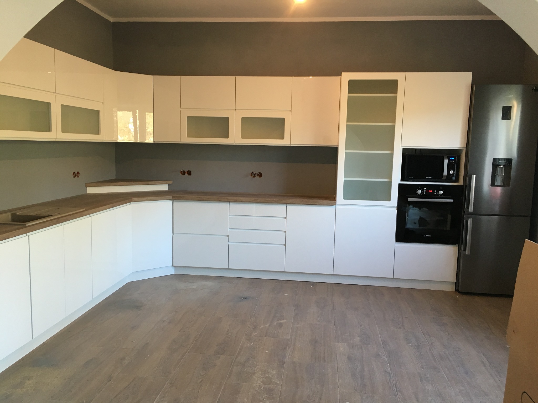 Biała, nowoczesna zabudowa kuchenna z drewnianym blatem, obejmująca szafki wiszące z przeszklonymi frontami, piekarnik, mikrofalówkę i lodówkę, na tle szarej ściany i drewnianej podłogi.