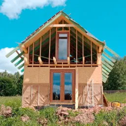 Surowy stan budynku w trakcie budowy z widocznymi drewnianymi elementami konstrukcyjnymi, oknami PCV z brązowymi ramami, białą drabiną opartą o ścianę i otoczeniem w postaci trawy i drzew.