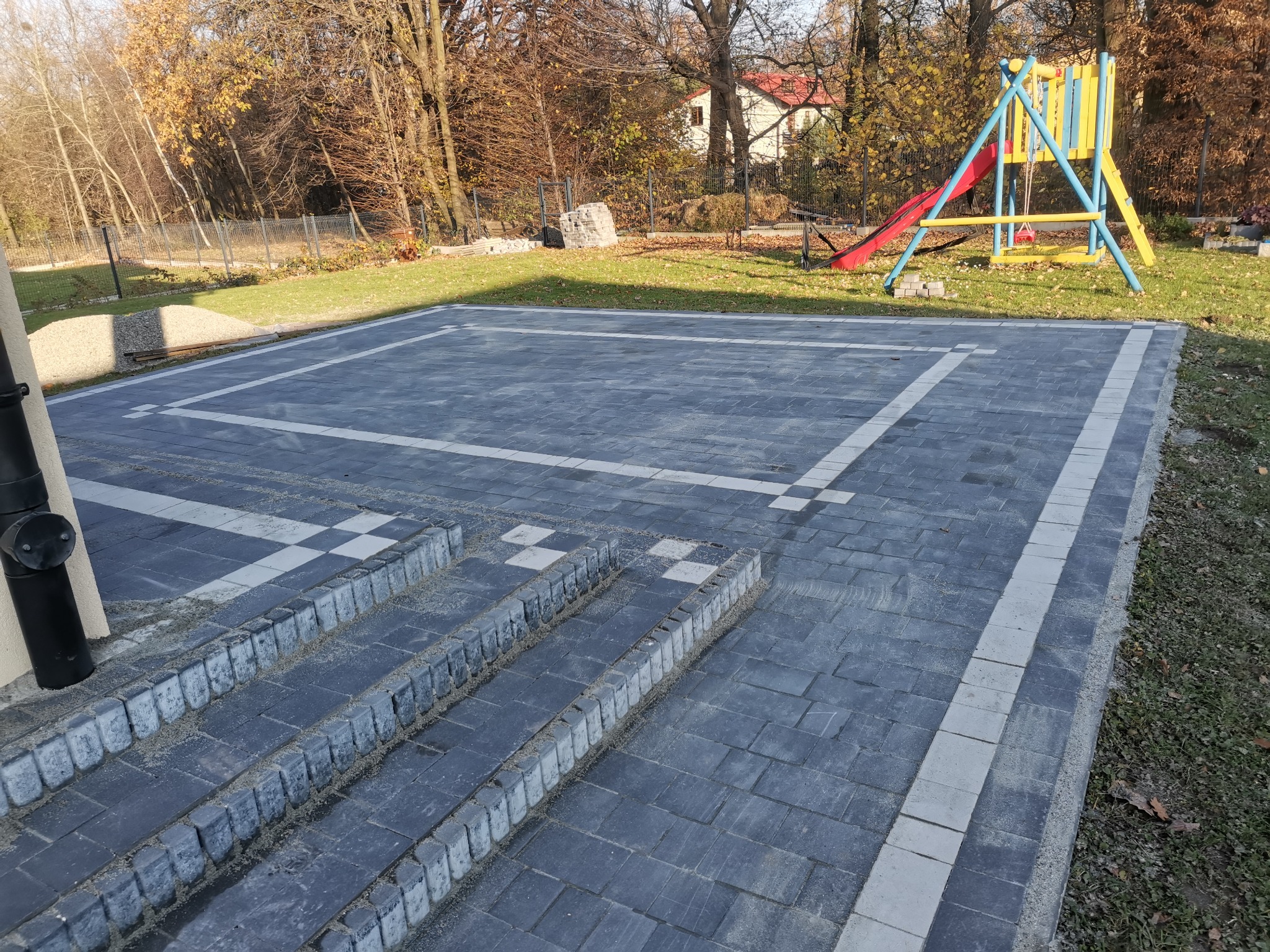Wykonana z ciemnoszarej kostki brukowej nawierzchnia z jasnymi obramowaniami i schodami, plac zabaw w tle, jesienna sceneria.
