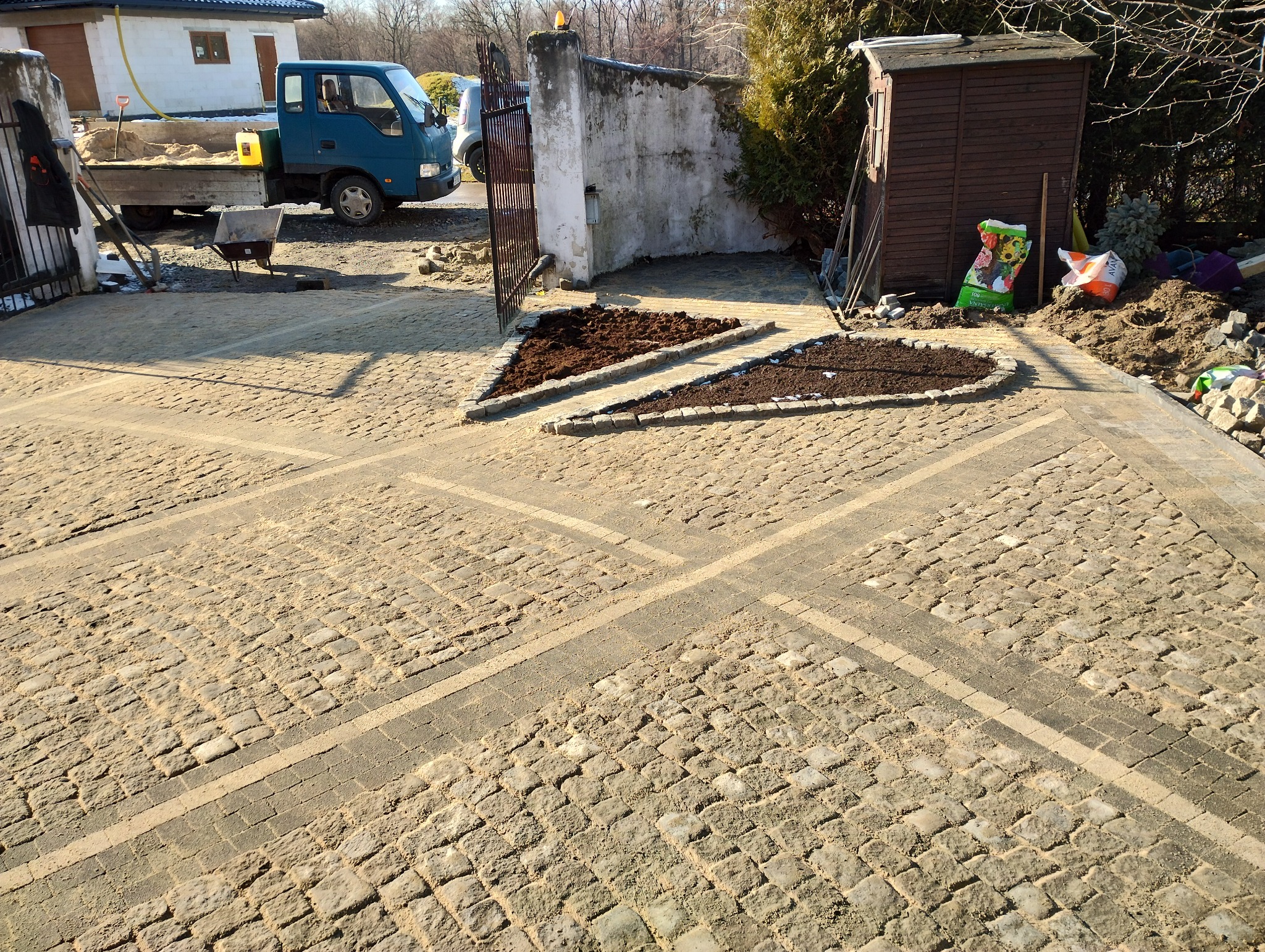 Ułożona kostka brukowa w kształcie rombów z wyznaczonymi miejscami na rabaty w kształcie trójkąta i owalu, w tle samochód dostawczy i domek narzędziowy.