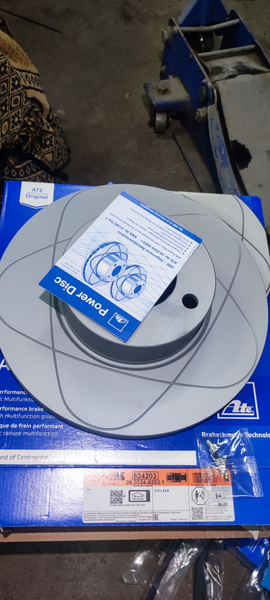 Nowe tarcze hamulcowe ATE Power Disc w oryginalnym opakowaniu, widoczna ulotka informacyjna i śruby montażowe, w tle podnośnik samochodowy.