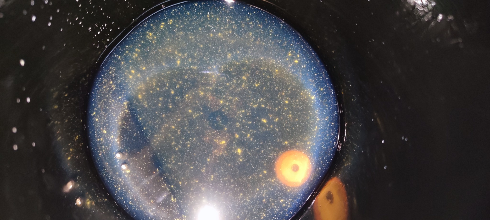 Pojemnik z płynem chłodniczym o niebieskawym odcieniu i złotymi drobinkami, widoczne refleksy światła i zabrudzenia na ściankach naczynia.