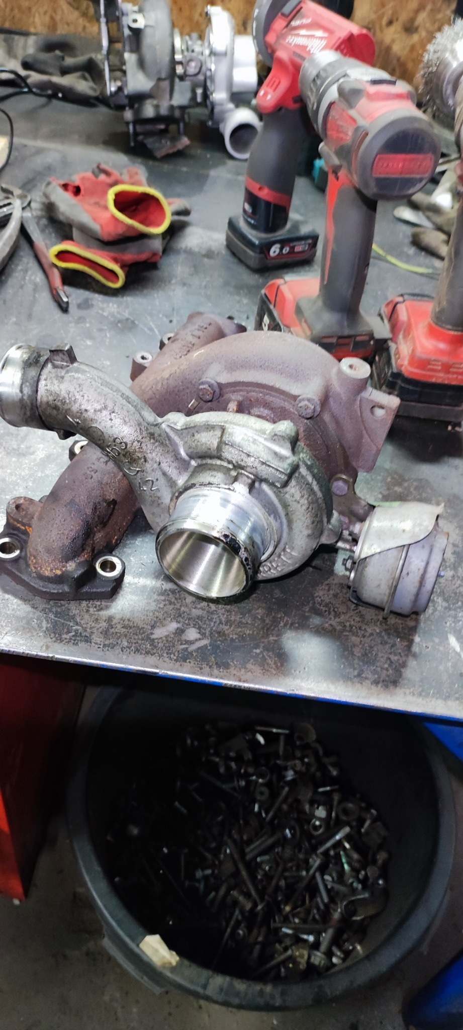 Używana turbosprężarka samochodowa z widocznymi śladami eksploatacji, leżąca na metalowym stole warsztatowym obok narzędzi akumulatorowych i pojemnika ze śrubami.