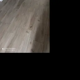 Ułożona pod kątem podłoga z paneli imitujących drewno w odcieniach szarości, widoczny fragment białej szafki w rogu.