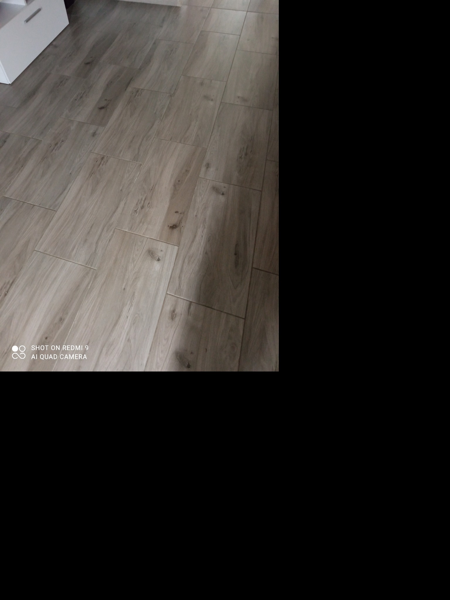 Ułożona pod kątem podłoga z paneli imitujących drewno w odcieniach szarości, widoczny fragment białej szafki w rogu.