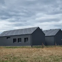 Dwa nowoczesne, pomalowane na ciemnoszaro domy jednorodzinne z szarą dachówką, widoczne zza suchej trawy na tle pochmurnego nieba.