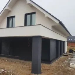 Nowoczesny dom jednorodzinny z jasną elewacją i ciemnymi elementami wykończeniowymi, widoczne czarne rolety zewnętrzne i kolumna podtrzymująca, teren wok&oacute;ł domu w trakcie prac wykończeniowych.