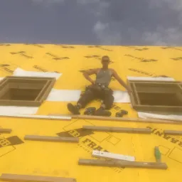 Pracownik w okularach przeciwsłonecznych siedzi na żółtej membranie dachowej z dwoma drewnianymi otworami okiennymi, obok leżą narzędzia i kawałki drewna.