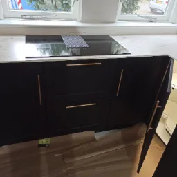 Ciemne szafki kuchenne z marmurowym blatem i płytą indukcyjną, otwarte narożne szafki z półkami obrotowymi, widoczne panele podłogowe.