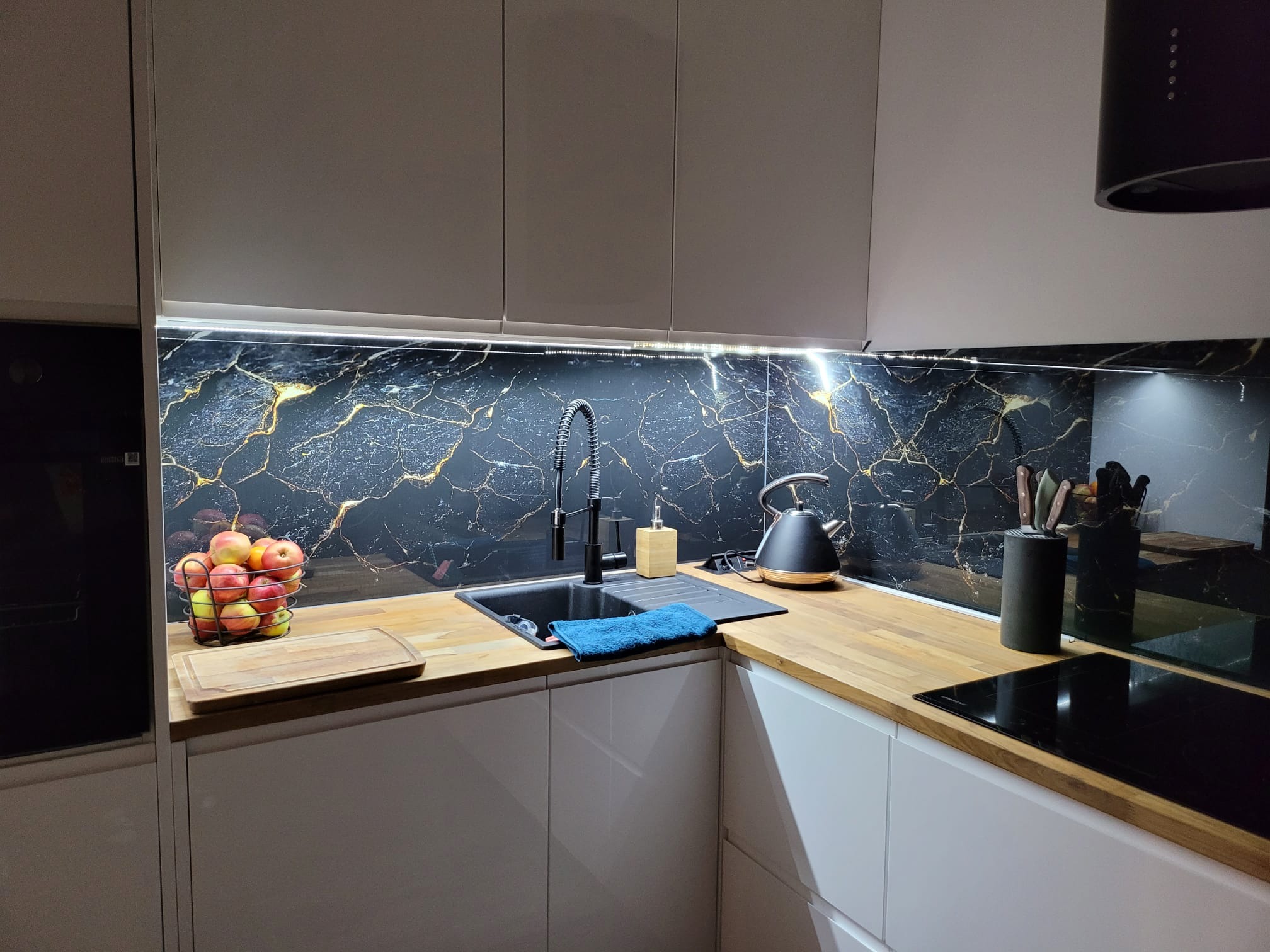 Nowoczesna kuchnia z białymi szafkami, blatem z jasnego drewna i czarnym panelem ściennym z marmurowym wzorem, oświetlona listwą LED pod szafkami górnymi, z czarnym zlewem i baterią.