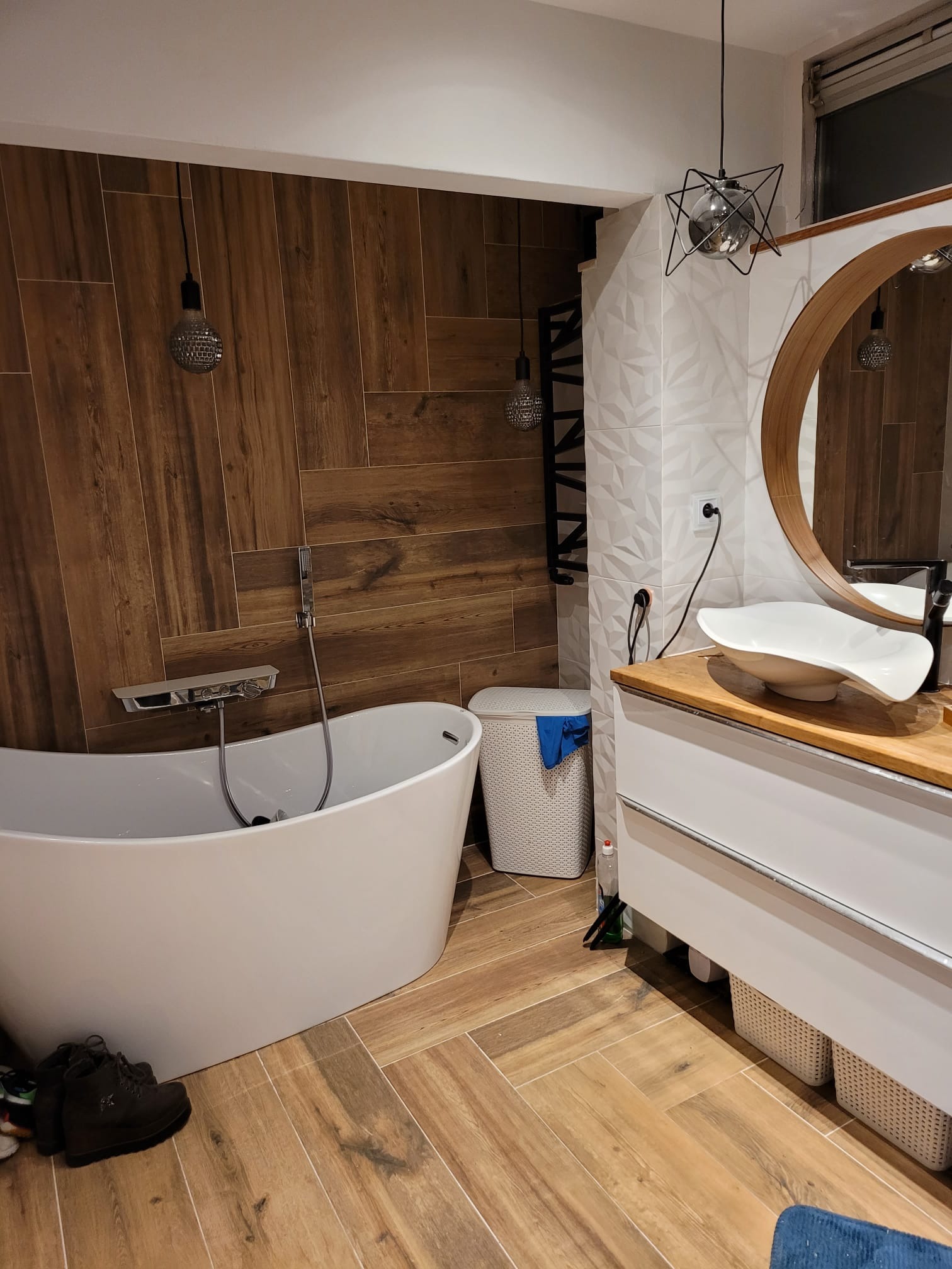 Nowoczesna łazienka z wolnostojącą wanną, drewnopodobnymi płytkami i okrągłym lustrem, oświetlona designerskimi lampami wiszącymi, z widocznymi butami obok wanny.