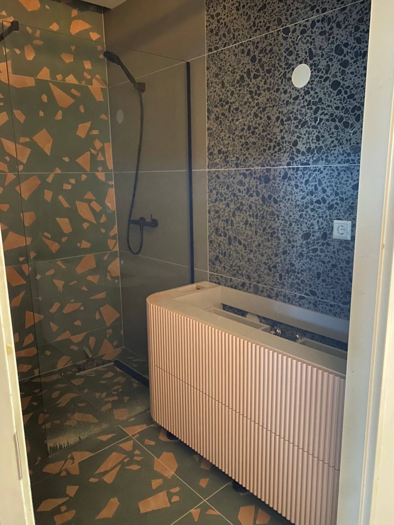 Nowoczesna łazienka z kabiną prysznicową, umywalką z szafką o ryflowanych frontach w kolorze pudrowego różu oraz płytkami w geometryczne wzory na ścianach i podłodze.