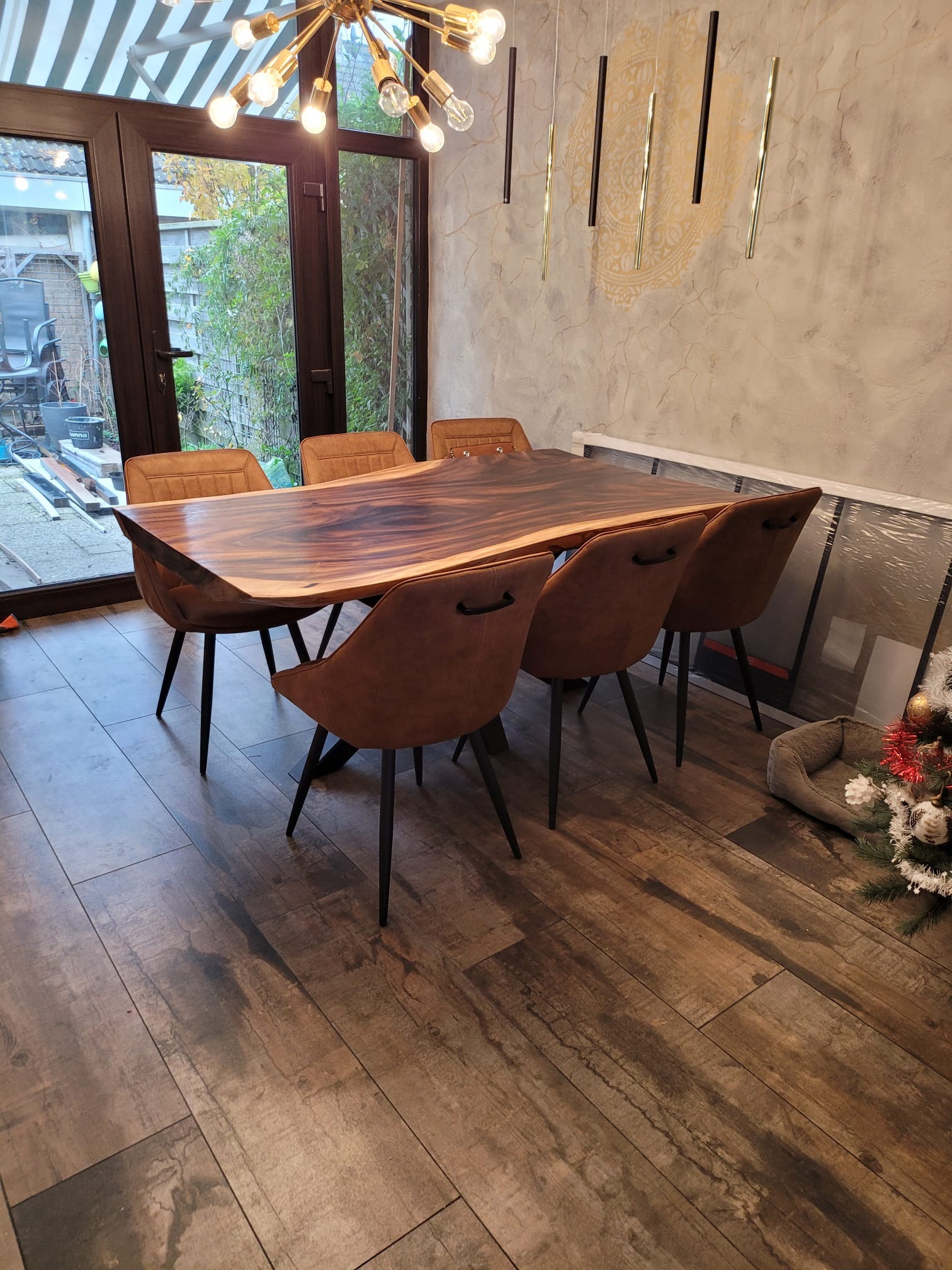 Jadalnia z dużym, drewnianym stołem o nieregularnych krawędziach, otoczonym sześcioma brązowymi krzesłami na tle ściany z dekoracyjnym wzorem i okna z widokiem na ogród.