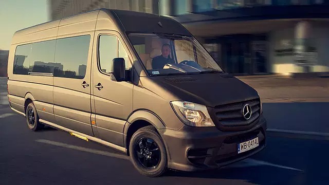 Szaro-brązowy Mercedes Sprinter z kierowcą, widoczny z przodu pod kątem, jadący po asfalcie w miejskiej scenerii.